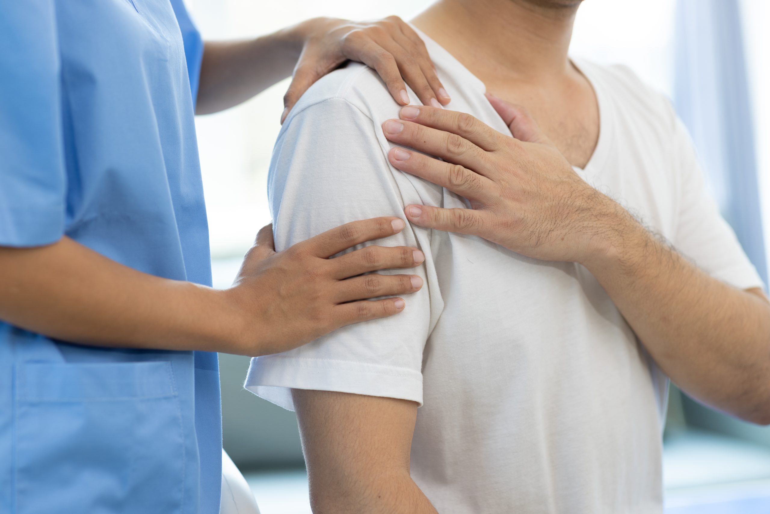 Doctor diagnosing man's shoulder pain in hospital examination room. Nurse doing massage and physiotherapy to treat patient's shoulder.