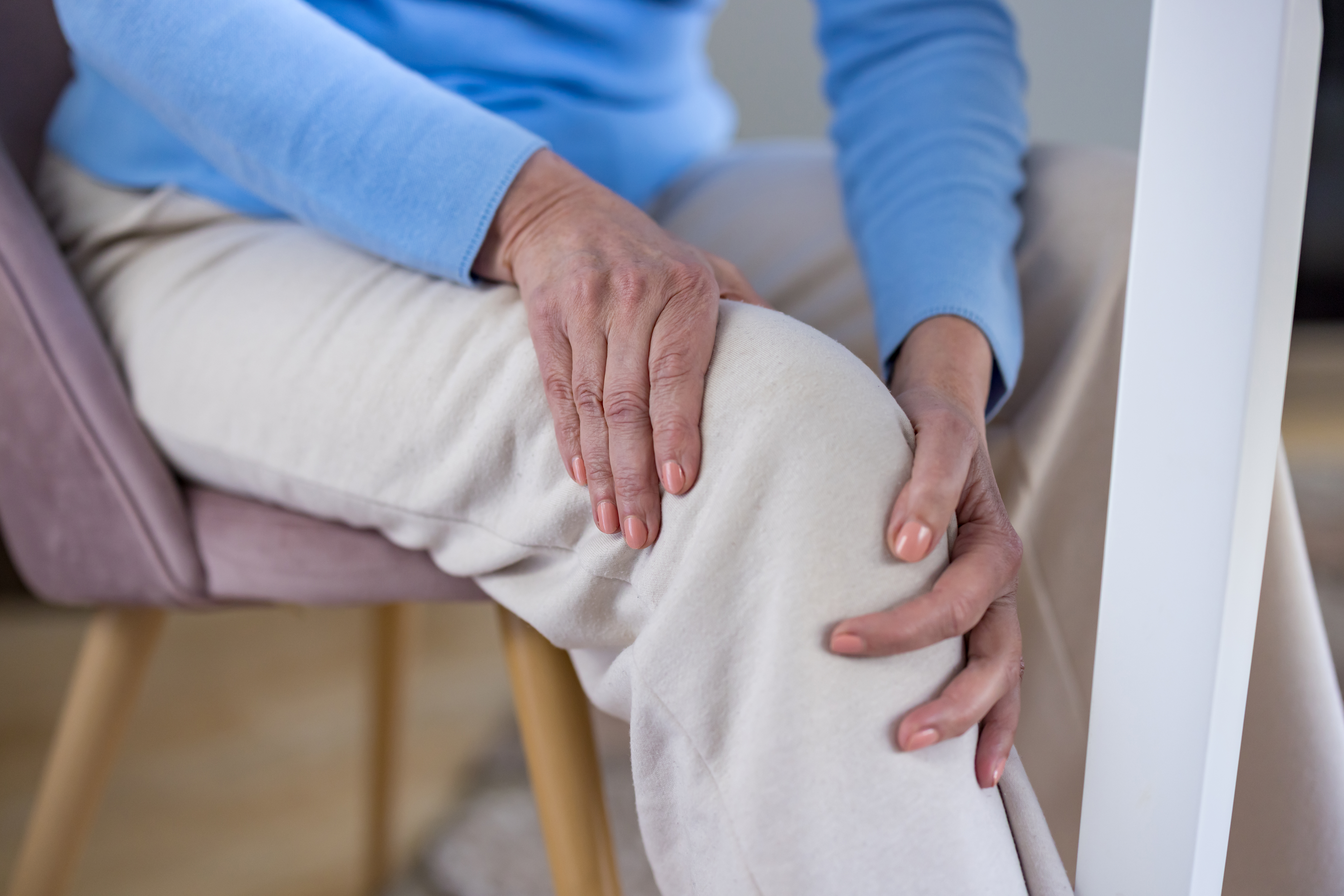 Senior woman sitting on a chair holding her painful knee with both hands, experiencing discomfort and health problems often associated with aging, arthritis, or injury